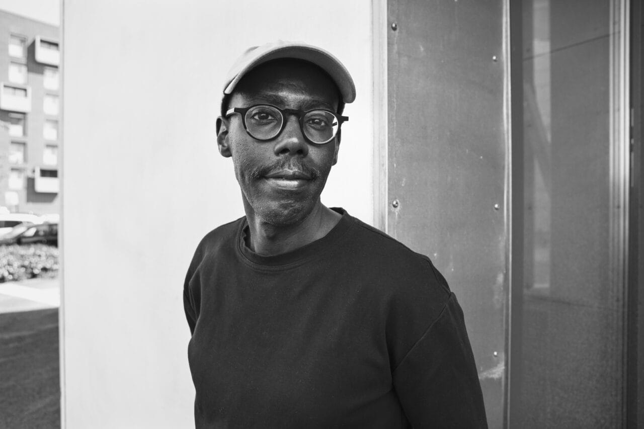 Mies silmälaseilla seisoo seinää vasten. Kuva on mustavalkoinen.