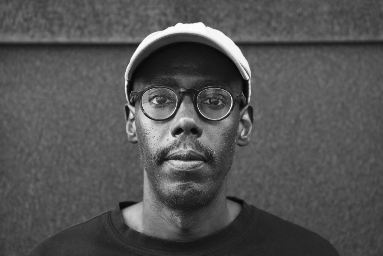 Mustavalkoinen muotokuva miehestä lippalakissa ja silmälaseissa katsomassa suoraan kameraan.