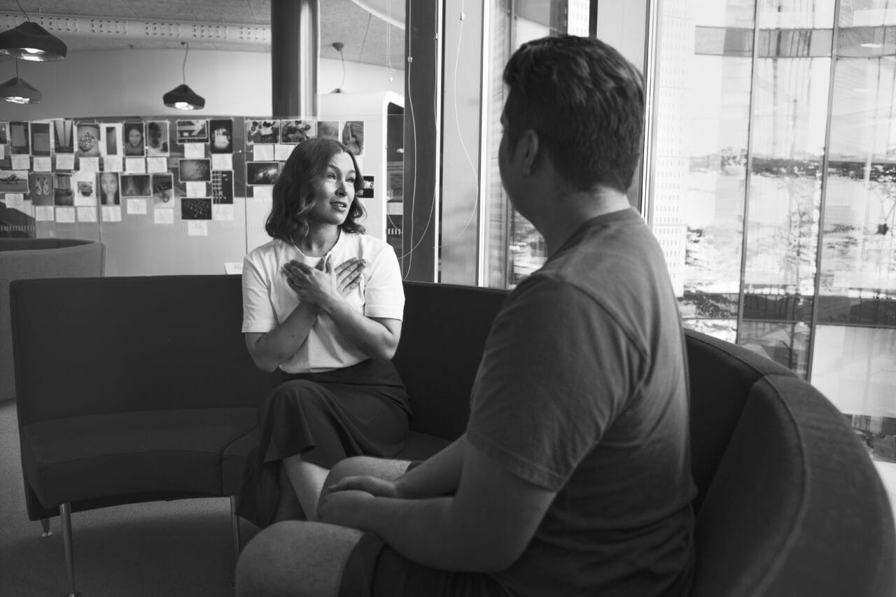 Kaksi henkilöä keskustelee sohvilla istuen sisätilassa. Kuva on mustavalkoinen.