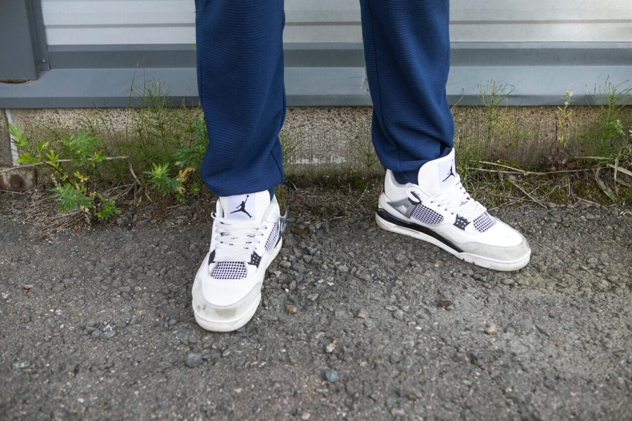 Kuva henkilön jaloista ja kuvassa korostuu henkilön pitämät valkoiset tennarit. Henkilö seisoo ulkona seisän edessä.