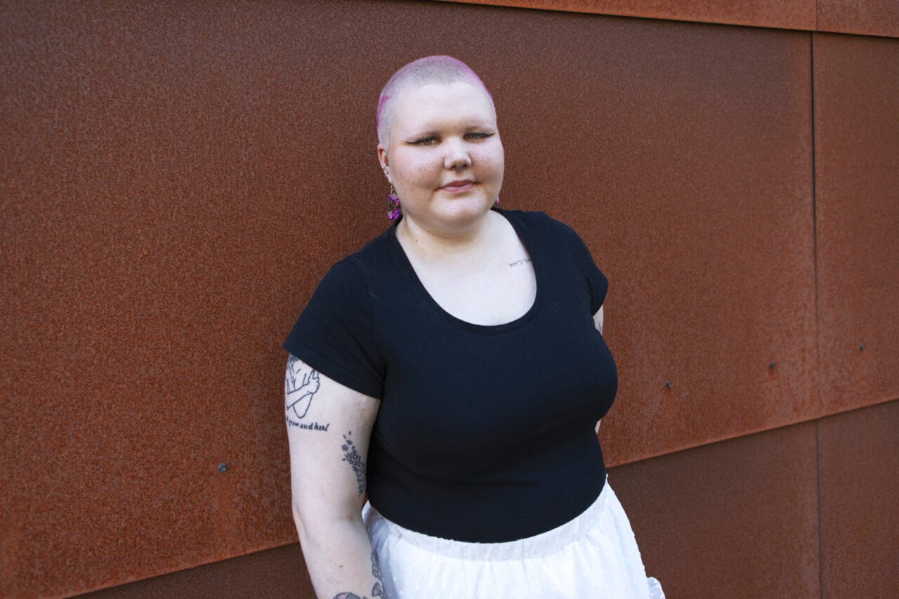 Henkilö seisoo ruskeaa seinää vasten ja katsoo kameraan.