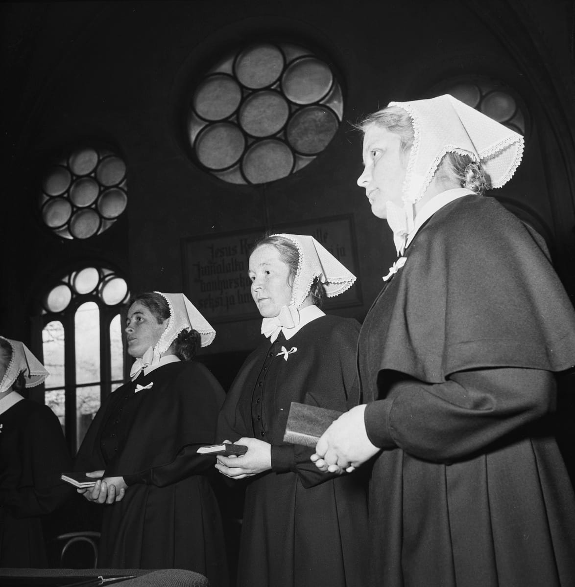 Vuonna 1954 Helsingissä Diakonissalaitoksen kirkossa otetussa mustavalkoisessa valokuvassa seisoo rivi diakonissoiksi vihittäviä naisia perinteisissä univormuissaan. Heillä on tummat viitat ja valkoiset päähineet, ja he pitelevät kädessään kirjoja. Taustalla näkyy kirkon koristeellisia pyöreitä lasi-ikkunoita ja hämärä sisätila.