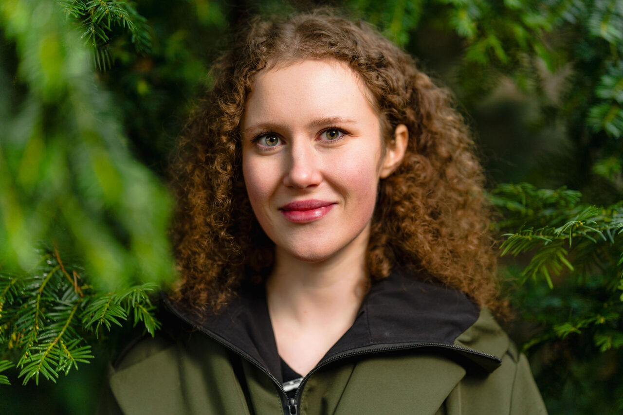 Kiharahiuksinen henkilö seisoo puiden edessä hymyillen.