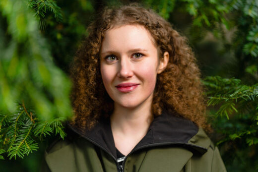 Kiharahiuksinen henkilö seisoo puiden edessä hymyillen.