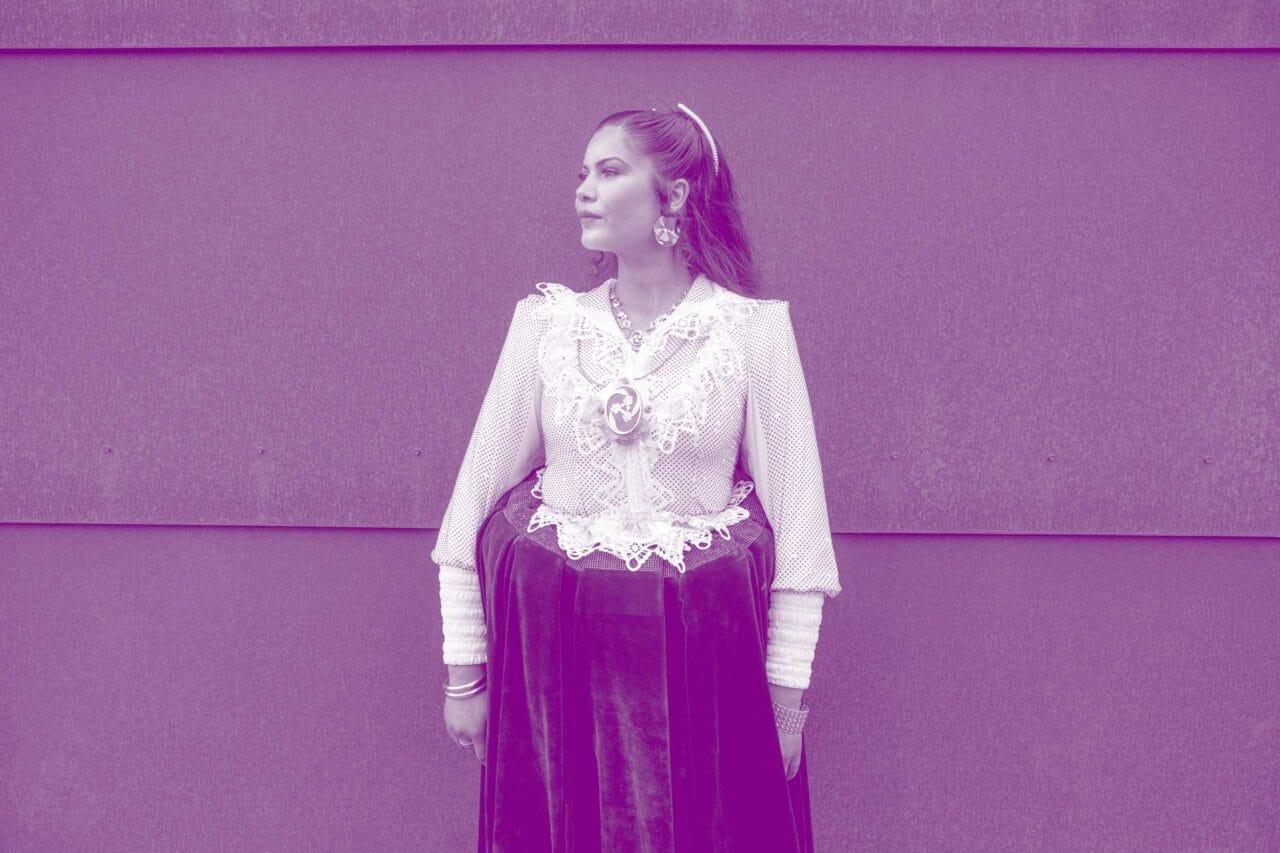 Nainen seisoo seinän edessä ja katsoo sivulle, yllään romaninaisen puku. Kuva on sävytetty violetiksi.