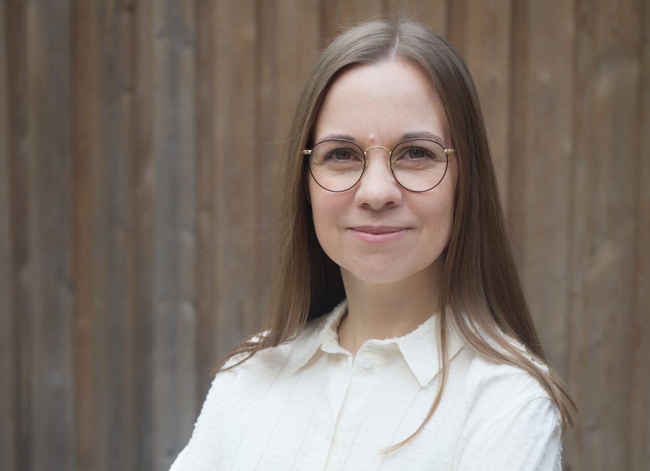 Muotokuva Aura Vuorenrinteestä, joka hymyilee silmälasit päässä vaaleassa paidassa. Taustalla on puuseinä.