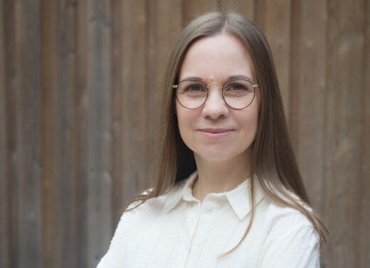 Muotokuva Aura Vuorenrinteestä, joka hymyilee silmälasit päässä vaaleassa paidassa. Taustalla on puuseinä.