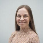 Lähikuva hymyilevästä Aura Vuorenrinteestä, jolla on silmälasit ja kuviollinen, korkeakauluksinen paita. Tausta on harmaa.