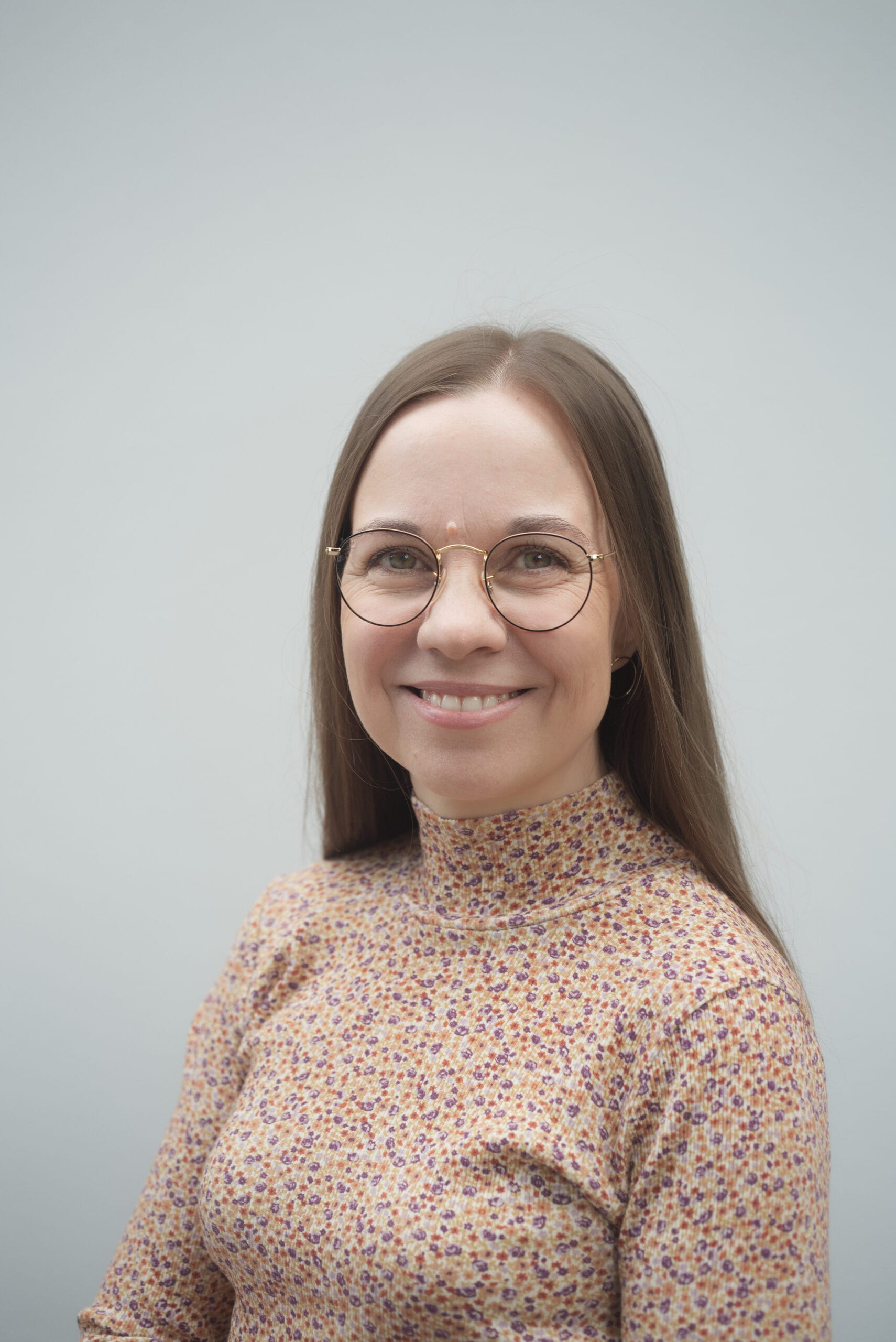Lähikuva hymyilevästä Aura Vuorenrinteestä, jolla on silmälasit ja kuviollinen, korkeakauluksinen paita. Tausta on harmaa.