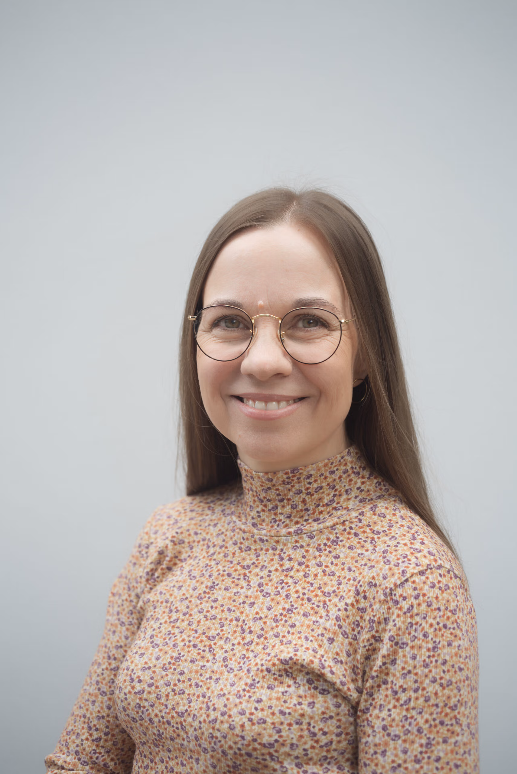 Lähikuva hymyilevästä Aura Vuorenrinteestä, jolla on silmälasit ja kuviollinen, korkeakauluksinen paita. Tausta on harmaa.