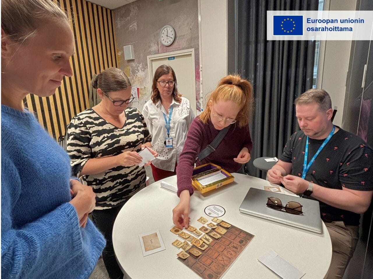 Viisi henkilöä seisoo ja istuu pöydän ympärillä pelaamassa korttipeliä. Pöydällä on pelikortteja ja pelilauta. Kuvan oikeassa yläkulmassa on Euroopan unionin osarahoittama -tunnus.