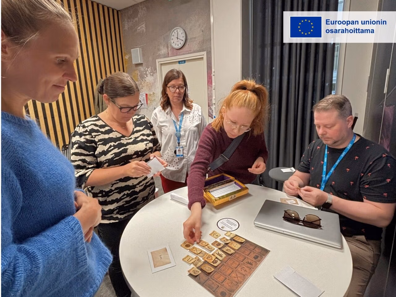 Viisi henkilöä seisoo ja istuu pöydän ympärillä pelaamassa korttipeliä. Pöydällä on pelikortteja ja pelilauta. Kuvan oikeassa yläkulmassa on Euroopan unionin osarahoittama -tunnus.
